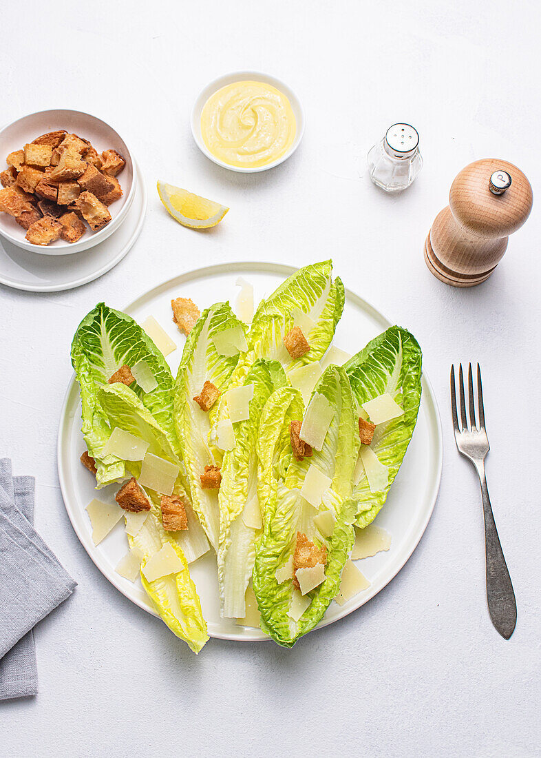 Draufsicht auf einen Caesar-Salat mit knackigem Kopfsalat, goldenen Croutons und gehobeltem Parmesan auf einem weißen Teller. Salz, Pfeffer, eine hölzerne Mühle, eine Crouton-Schale, eine Zitronenspalte und eine Schale mit Dressing sind ebenfalls zu sehen.