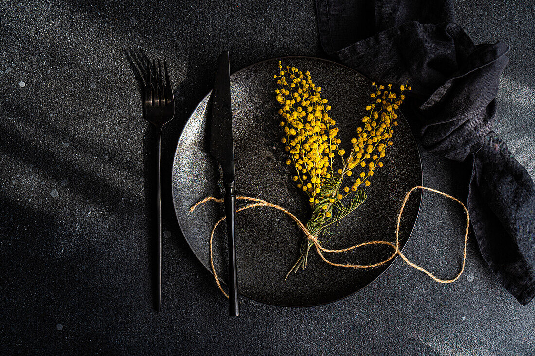 Top view of sophisticated black dinner plate and cutlery set adorned with bright yellow mimosa blossoms, tied with twine, on a dark textured background.