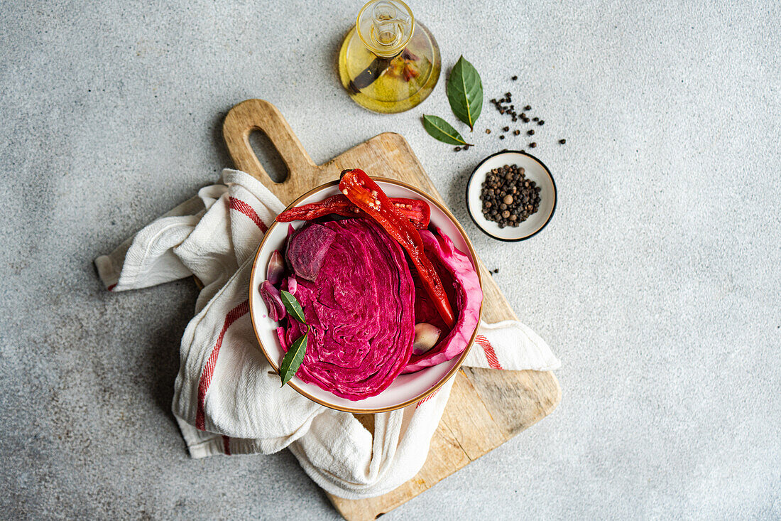 Vibrant beetroot fermented cabbage with spicy red peppers and garlic, served in a bowl on a rustic wooden cutting board, accompanied by olive oil, bay leaves, and whole peppercorns.