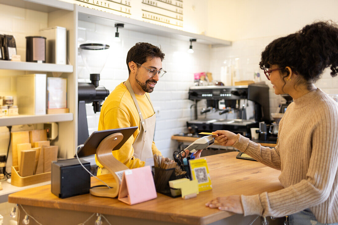 Ein lächelnder männlicher Barista im gelben Hemd nimmt die Zahlung einer Kundin an einem Kartenlesegerät in einem modernen Café entgegen.