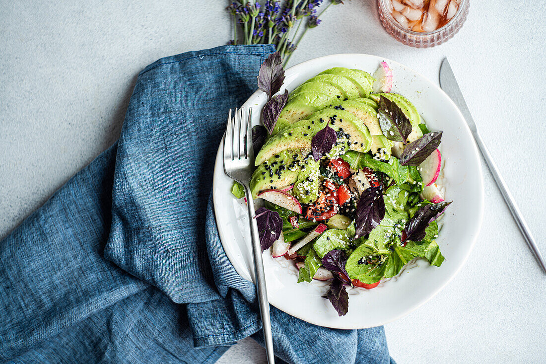 Bunter Gemüsesalat mit Kopfsalat, Oliven, Kirschtomaten, Gurkenscheiben, Radieschen, rotem Basilikum, bestreut mit weißem und schwarzem Sesam, kunstvoll präsentiert in einer weißen Schale mit blauer Serviette.