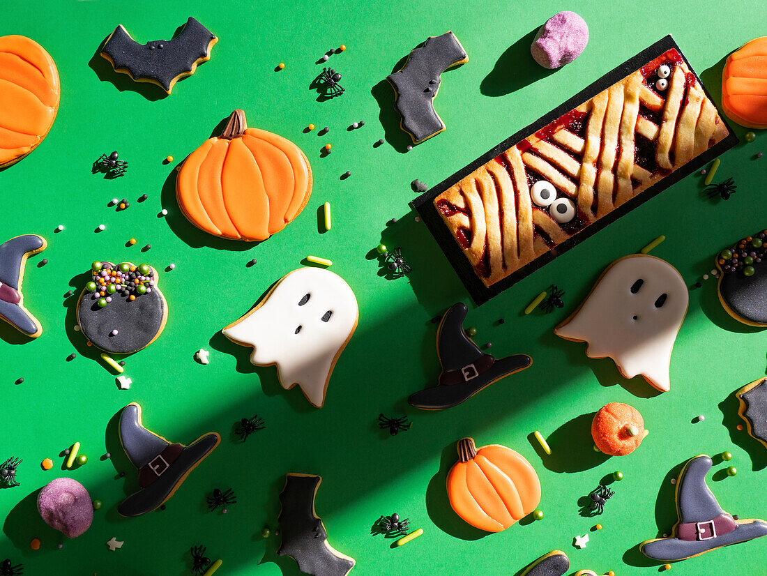 An assortment of homemade Halloween-themed cookies and sprinkles creatively arranged on a bright green background, with shapes such as ghosts, pumpkins, and witch hats