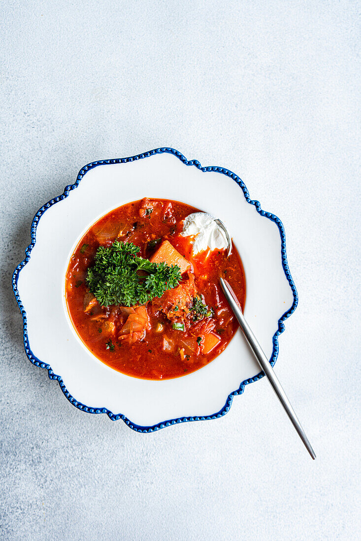 Eine Draufsicht auf eine traditionelle ukrainische Rote-Bete-Suppe, serviert mit einem Klecks saurer Sahne und garniert mit Petersilie, präsentiert auf einem verzierten weiß-blau umrandeten Teller