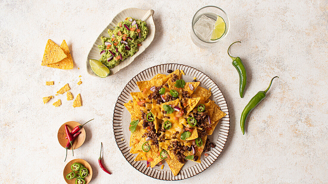 Top view of a delectable plate of nachos generously topped with cheese, minced meat, and fresh vegetables, alongside a serving of guacamole and a refreshing lime drink.