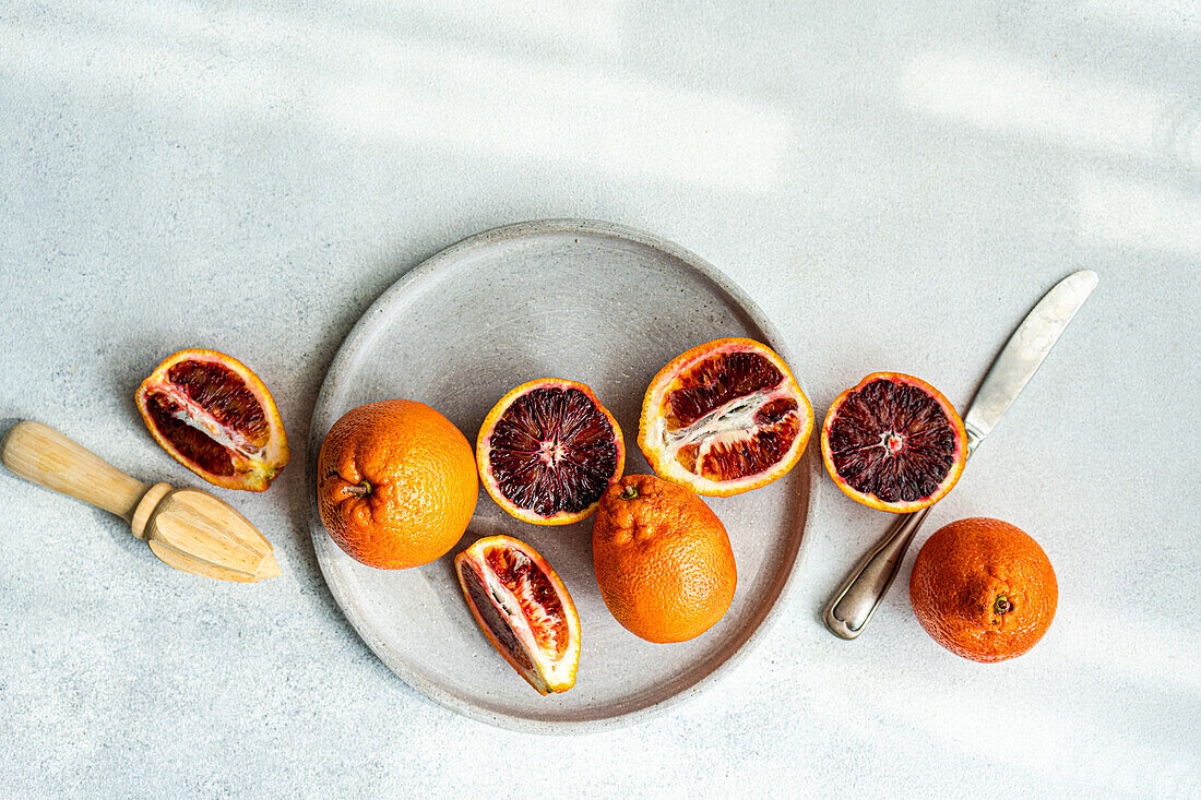 Vibrant blood oranges, both whole and sliced, arranged aesthetically on a gray concrete plate beside a wooden citrus reamer and stainless-steel knife