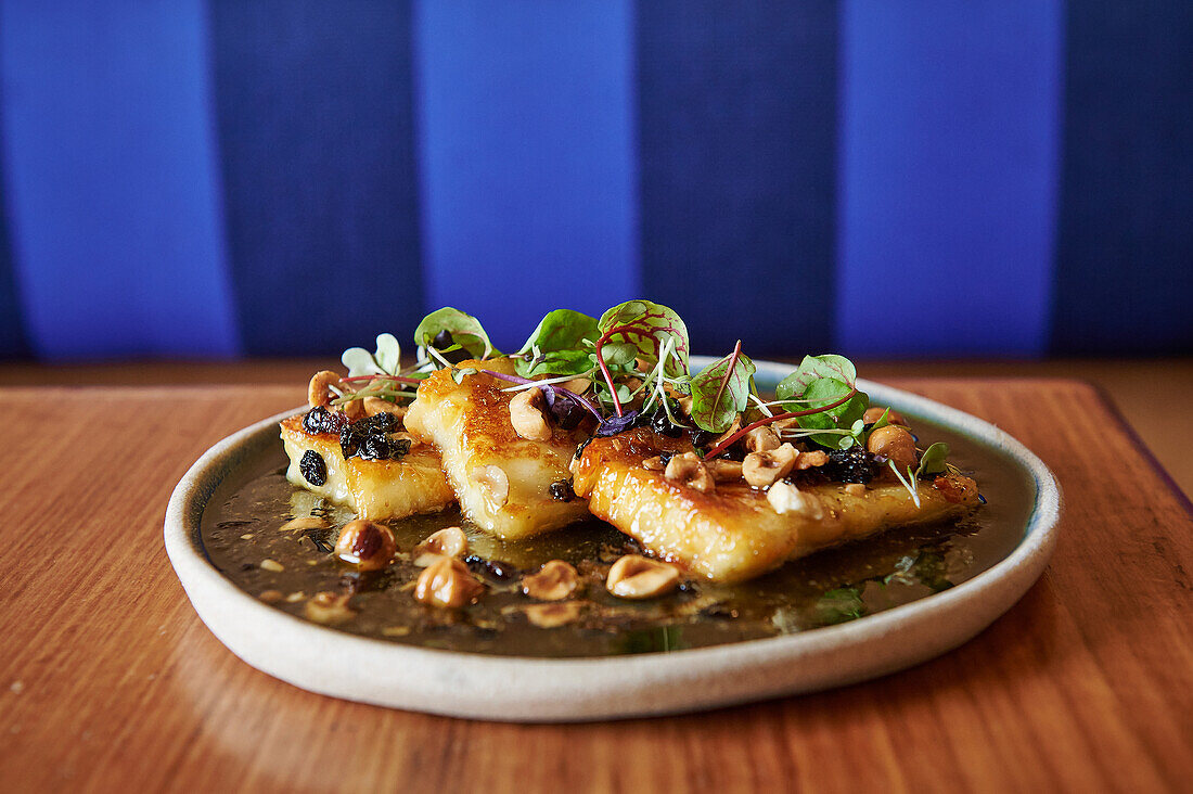 Dieses schmackhafte Gourmetgericht besteht aus goldbraunem Halloumi-Käse, garniert mit frischem Grün und knackigen Haselnüssen, präsentiert auf einem Keramikteller vor einem blau gepolsterten Hintergrund.