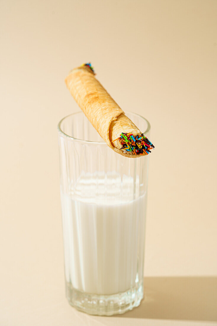 A freshly prepared waffle roll adorned with colorful sprinkles, perfectly balancing on a full glass of milk against a beige background.