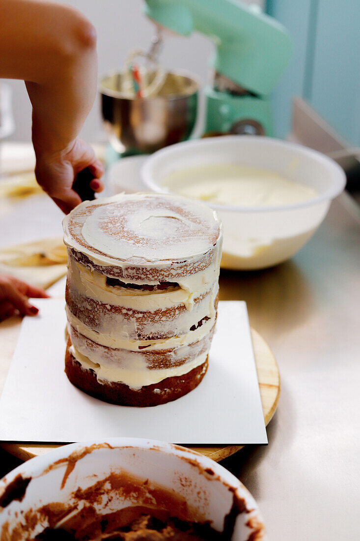 Eine unfertige Torte, die mit cremigem Zuckerguss überzogen wird, neben einer Rührschüssel und einem Küchenmixer im Hintergrund