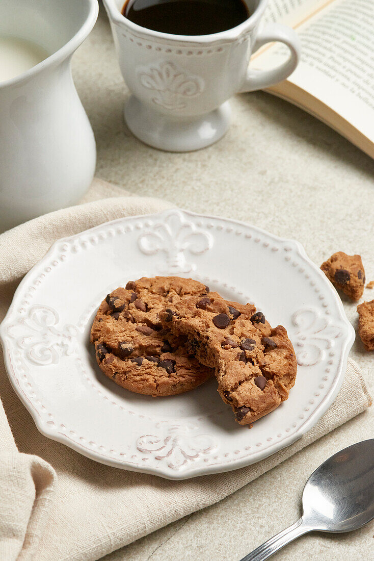 A comforting arrangement featuring a broken chocolate chip cookie on a decorative plate, with a cup of coffee and a book in the background, captured from above