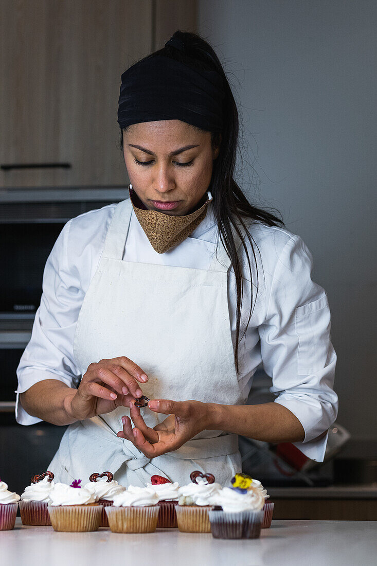 Focused female baker standing at counter in kitchen of bakery and decorating sweet vegan cupcakes with berries