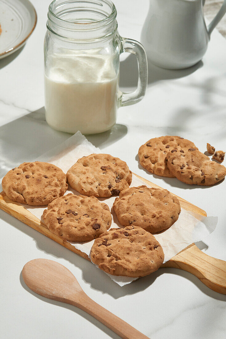 Sunlit kitchen scene with chocolate chip cookies on a wooden spatula beside a jar of milk and a ceramic pitcher