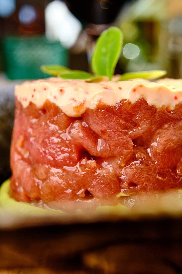 A close-up shot of a delicately layered tuna tartare topped with a creamy, spicy sauce and a green basil leaf, served on a natural stone platter.