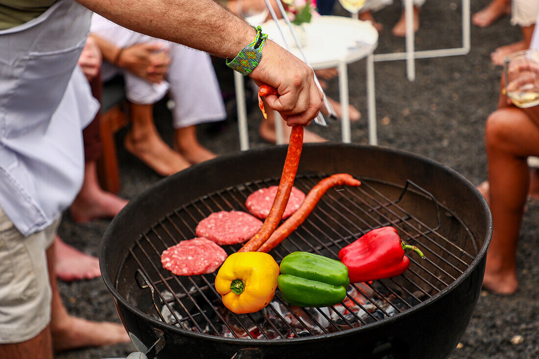 Cropped unrecognizable person grilling sausages and vegetables on a barbecue at an outdoor gathering. Background includes guests socializing, enjoying drinks and music.