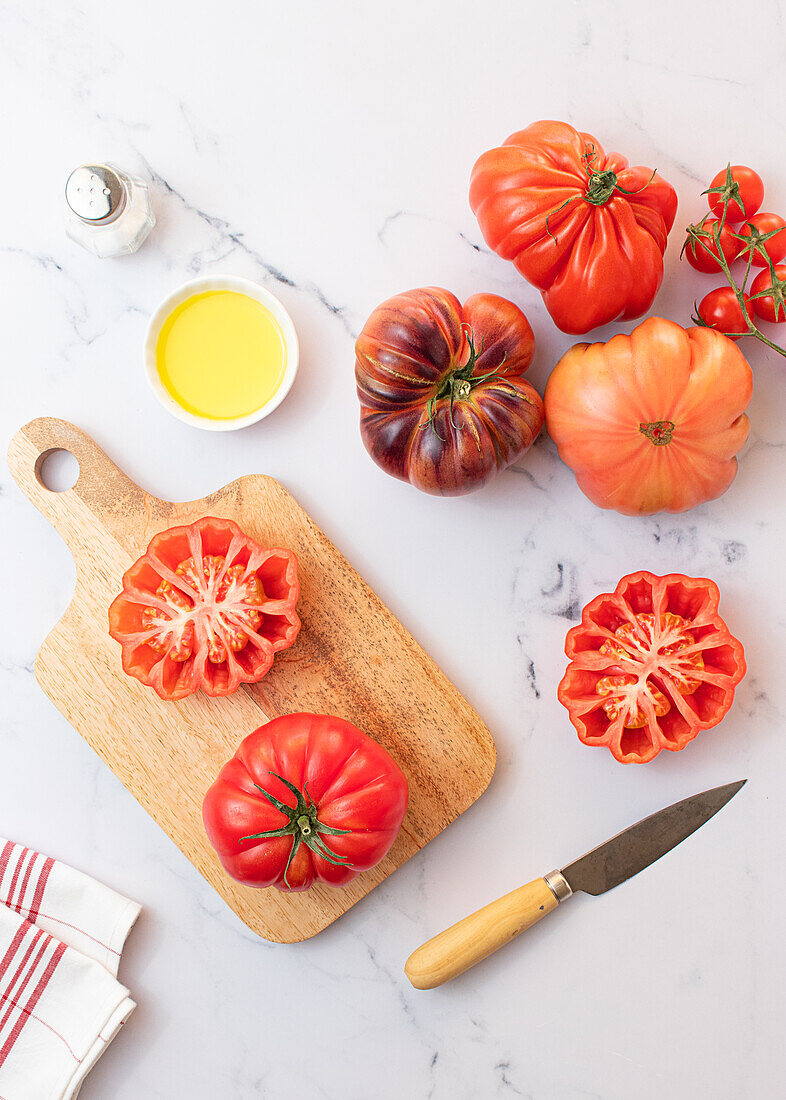 Von oben: Zubereitung eines Tomatensalats auf einem Holzbrett mit einem Messer, Salz, Olivenöl und einer Auswahl von Tomaten