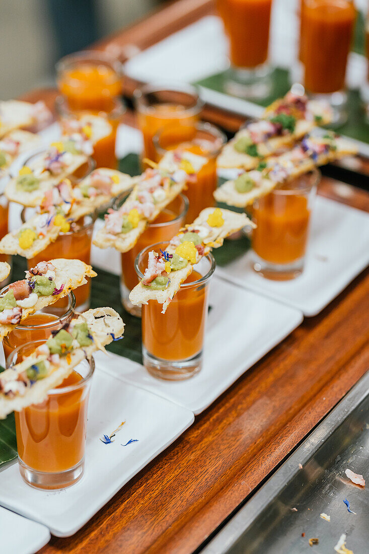 A sophisticated culinary setup featuring shot glasses of gazpacho soup garnished with exquisitely prepared gourmet toppings