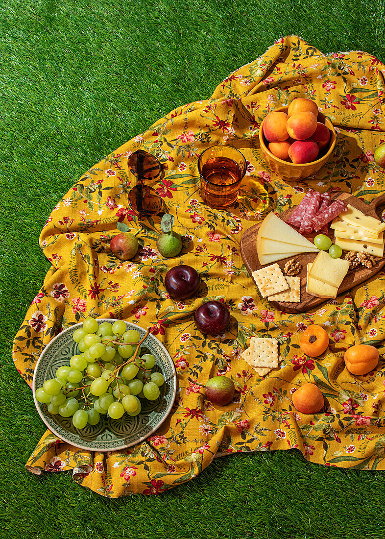 A beautifully arranged summer picnic set up on a floral yellow cloth, featuring a selection of fruits, cheeses, crackers, and meats, perfect for a sunny day outdoors