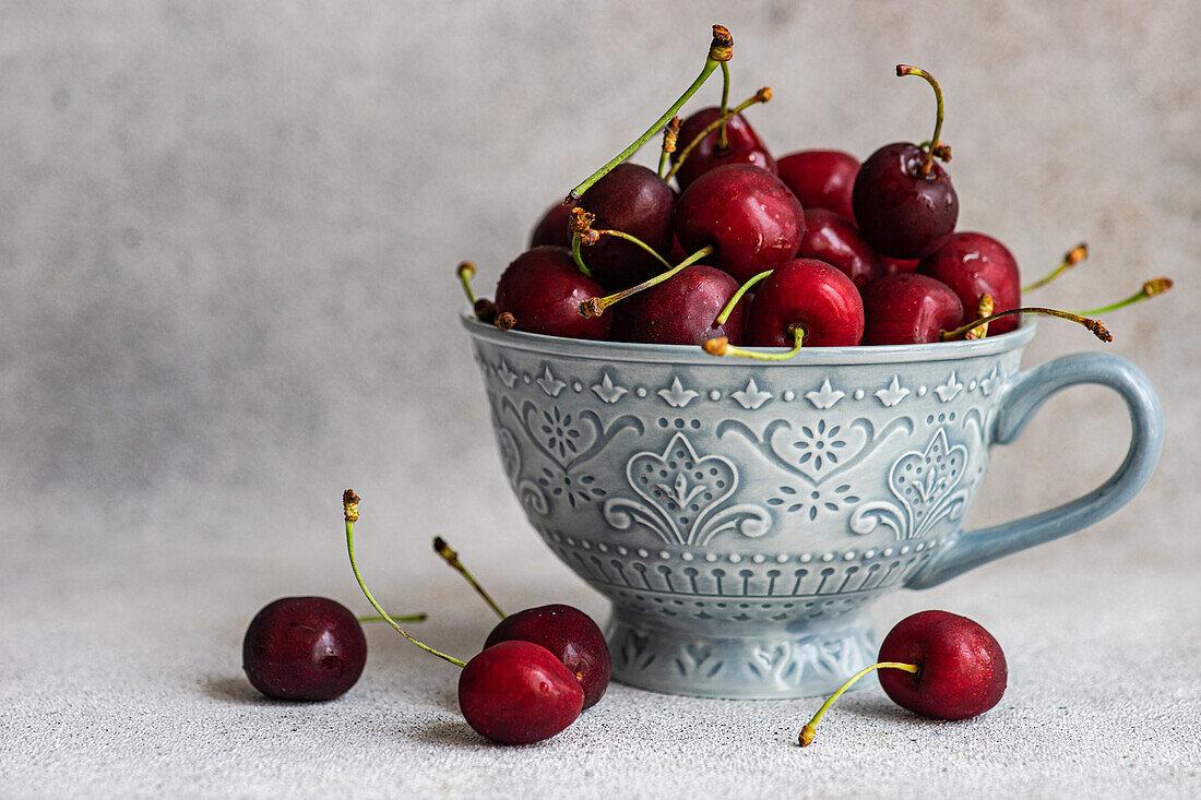 Decorative grey ceramic mug filled with juicy red cherries. The cherries overflow onto a textured grey surface, highlighting their vibrant color and fresh appearance.