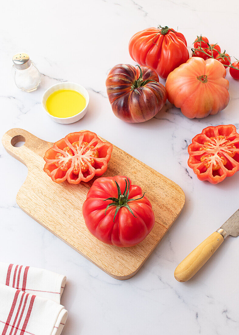 From above tomato salad preparation on wooden board with a knife, salt, olive oil and a selection of tomatoes