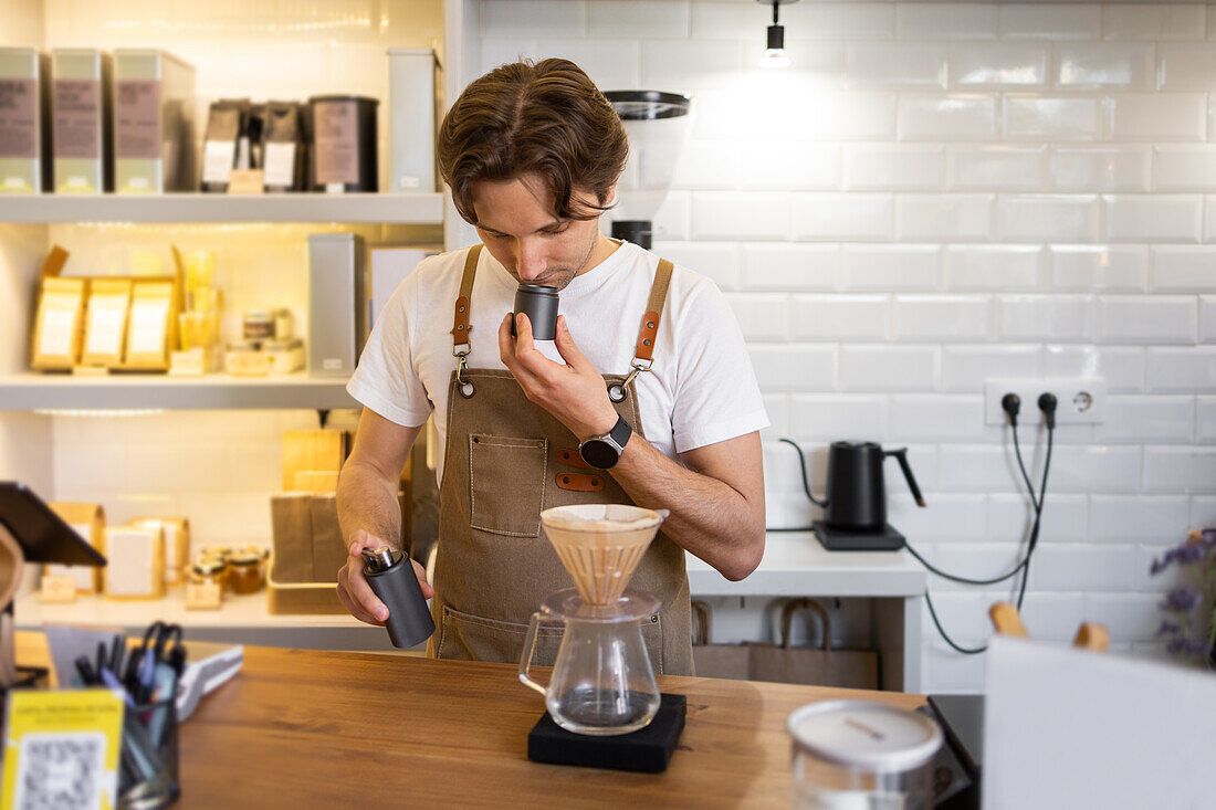 A focused barista in a canvas apron smells freshly ground beans while brewing coffee using a pour-over method in a bright, trendy cafe setting.