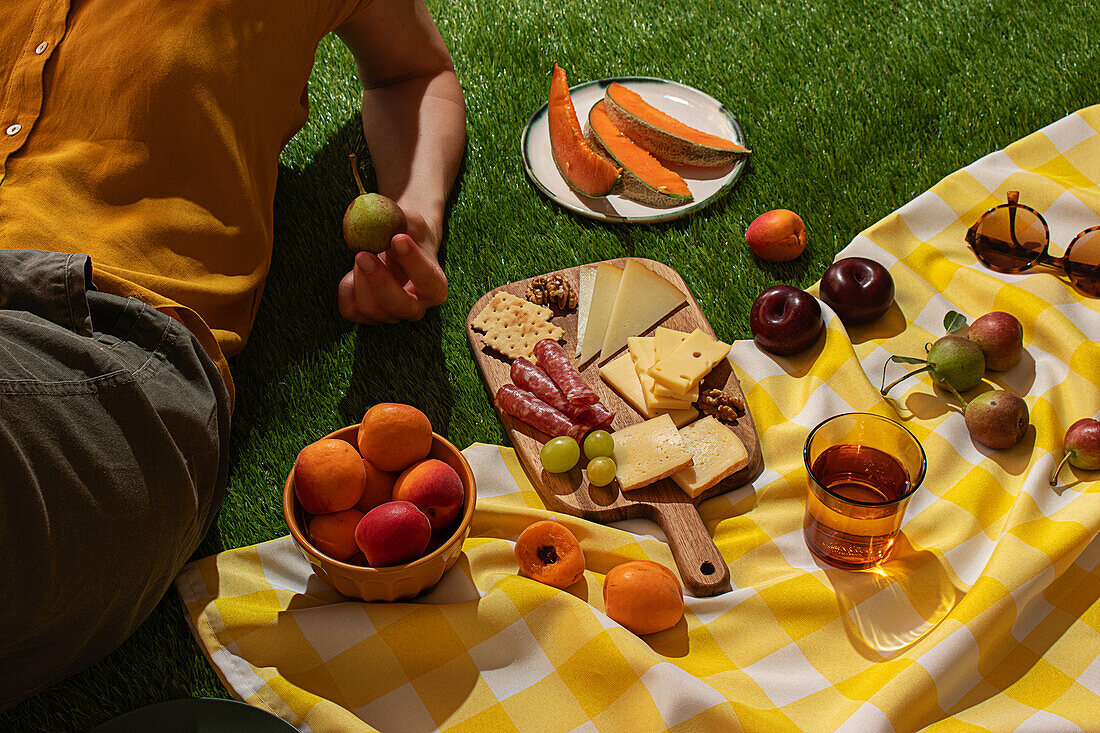 An anonymous person on a serene summer picnic, with a vibrant yellow and white blanket laden with a variety of cheeses, fruit and drinks in the natural daylight