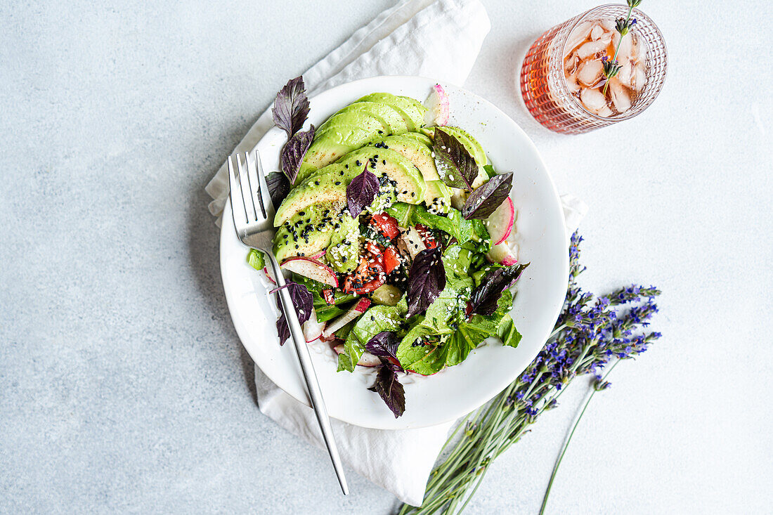 A vibrant bowl of vegetable salad with fresh lettuce, olives, juicy tomatoes, crisp cucumber, radishes, and red basil herb, sprinkled with both white and black sesame seeds, served alongside a refreshing iced drink and decorative lavender sprigs.