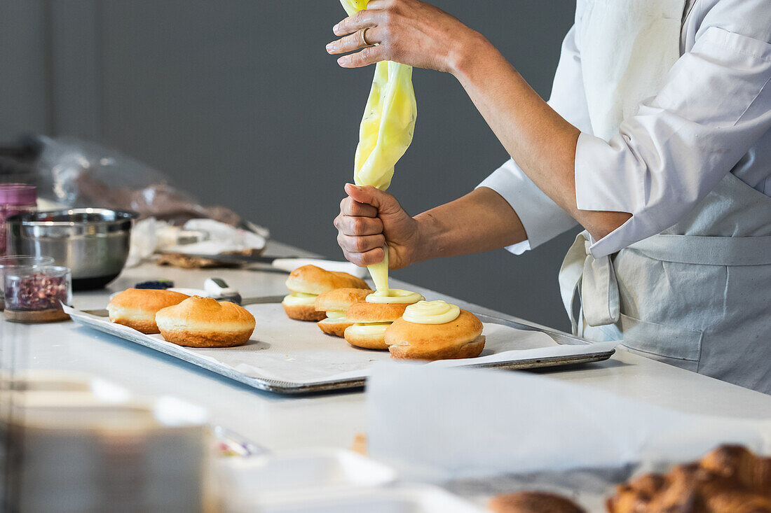 Seitenansicht eines anonymen Bäckers, der Sahne aus einem Spritzbeutel drückt, während er süße vegane Berliner in der Küche einer Bäckerei dekoriert