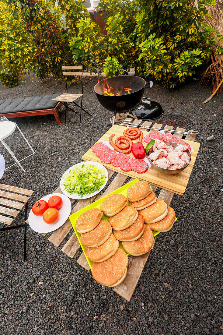 A backyard BBQ setting showing a grill with flames, surrounded by ingredients such as sausages, meats, salad, and buns set for preparation by friends enjoying music together.