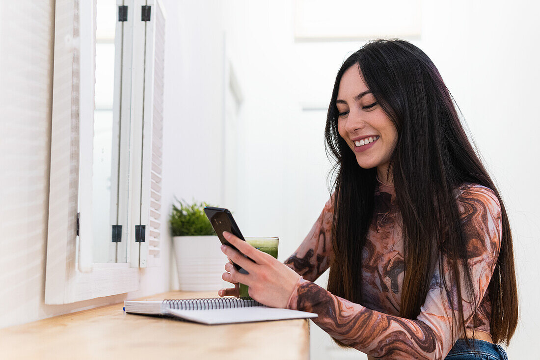 Smiling female sitting at counter with green smoothie in bar browsing on smartphone while writing in notepad