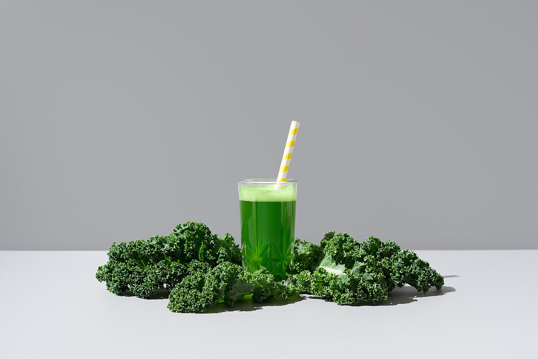 A vibrant green kale juice served in a transparent glass with a yellow-striped straw, accompanied by fresh kale leaves on a neutral background