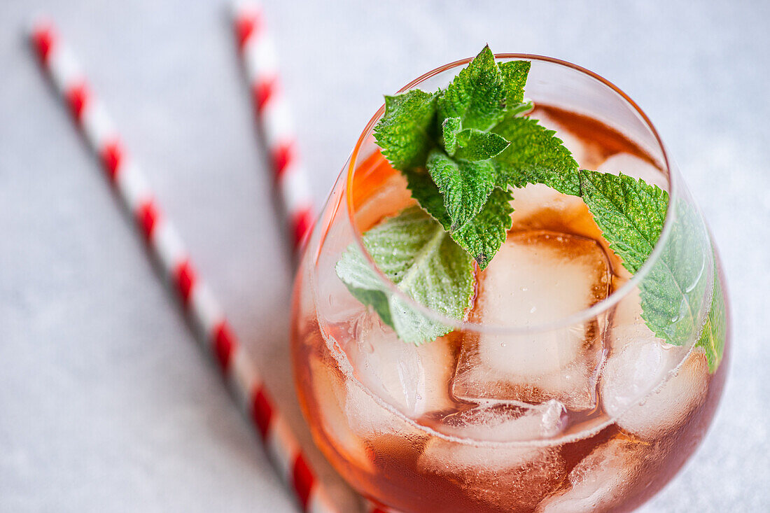 Nahaufnahme eines gekühlten Coca-Cola-Cocktails, garniert mit frischen Minzblättern in einem Glas, begleitet von rot-weiß gestreiften Strohhalmen, vor einem weichen grauen Hintergrund.