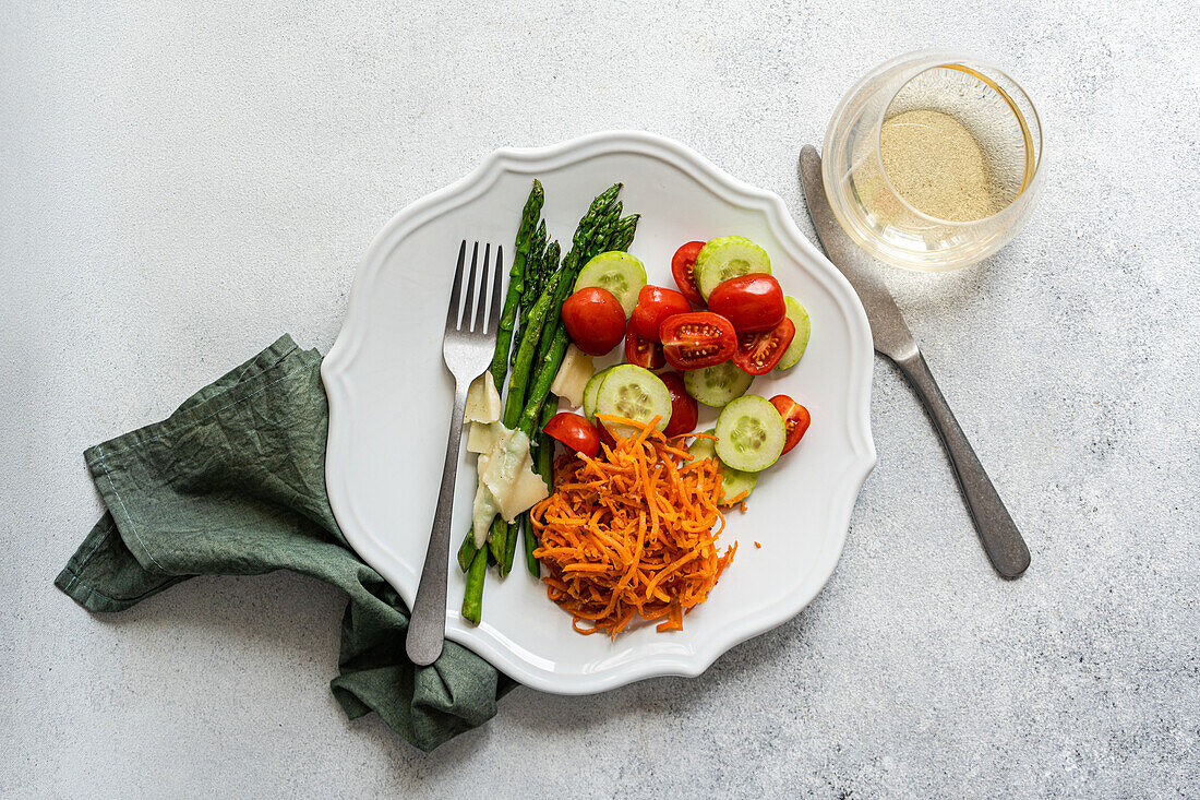 Draufsicht auf einen schön angerichteten Gemüseteller mit Spargelstangen, in Scheiben geschnittenen Tomaten und Gurken, geraspelten Karotten, Käse, neben einem Glas Wein und einer Schale mit rosa Salz.