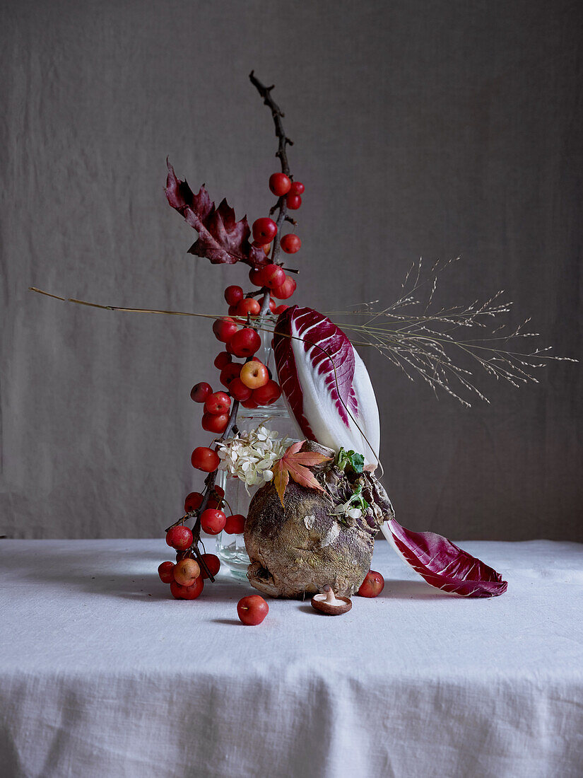 The image features a creative arrangement of tiny red crab apples, accompanied by a radicchio leaf and a swede, all elegantly displayed against a moody gray backdrop.