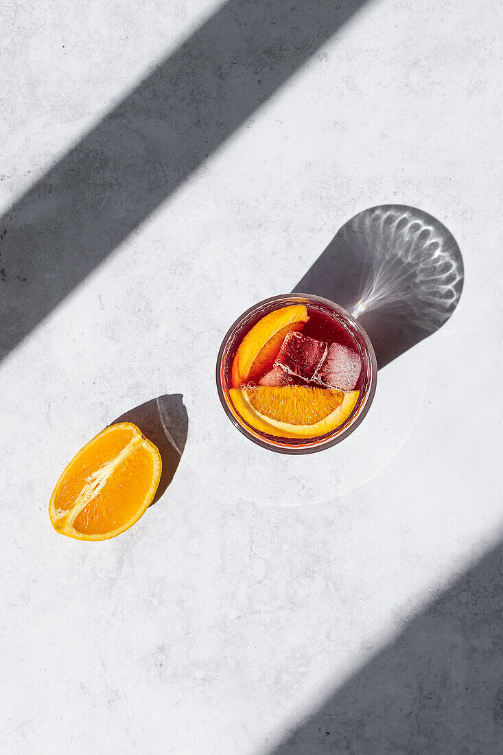 A vibrant glass of Tinto de Verano, garnished with a slice of orange, sits bathed in sunlight, casting a reflective shadow on a textured surface