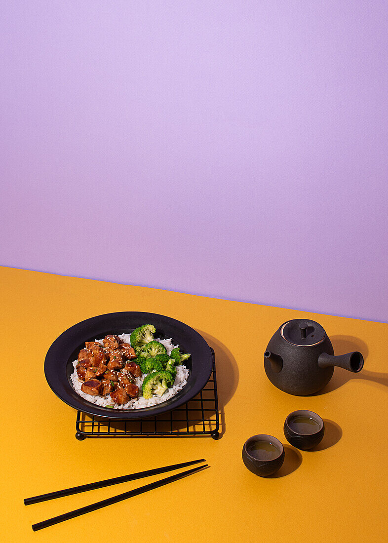 Artfully plated teriyaki chicken on a black plate, served with broccoli and rice, accompanied by a traditional tea set on a vibrant yellow and purple background.