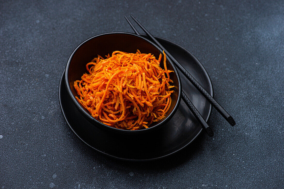 A black bowl filled with shredded carrot salad, accompanied by a pair of black chopsticks, presented on a textured dark gray surface