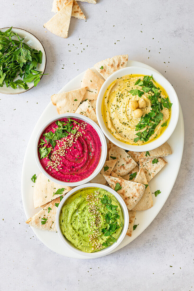 Top view three bowls of colorful homemade hummus—classic, beet, and avocado—served with pita bread and garnished with herbs.