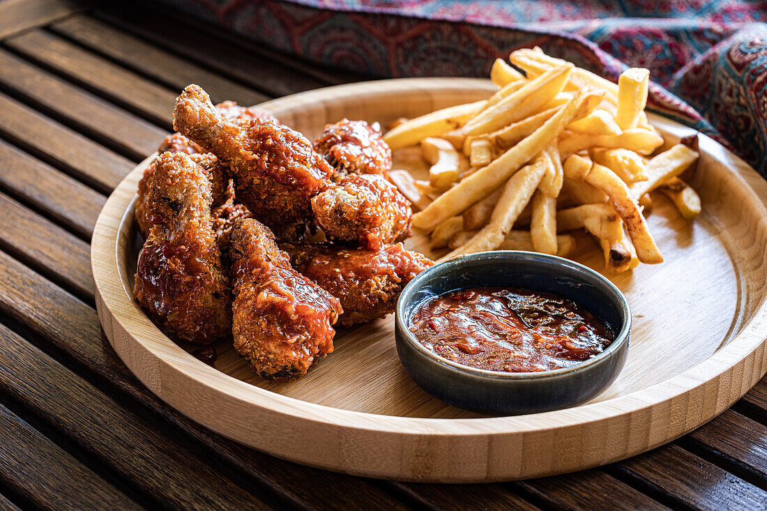 Crispy fried chicken hips served alongside golden french fries and a bowl of rich BBQ sauce, presented on a wooden plate with a rustic backdrop.