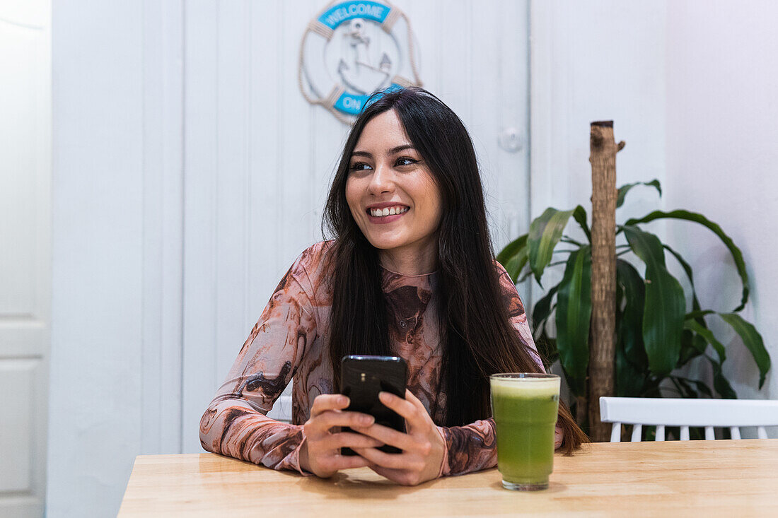 Positive female sitting at table with smoothie and browsing mobile phone while chilling at weekend in bar