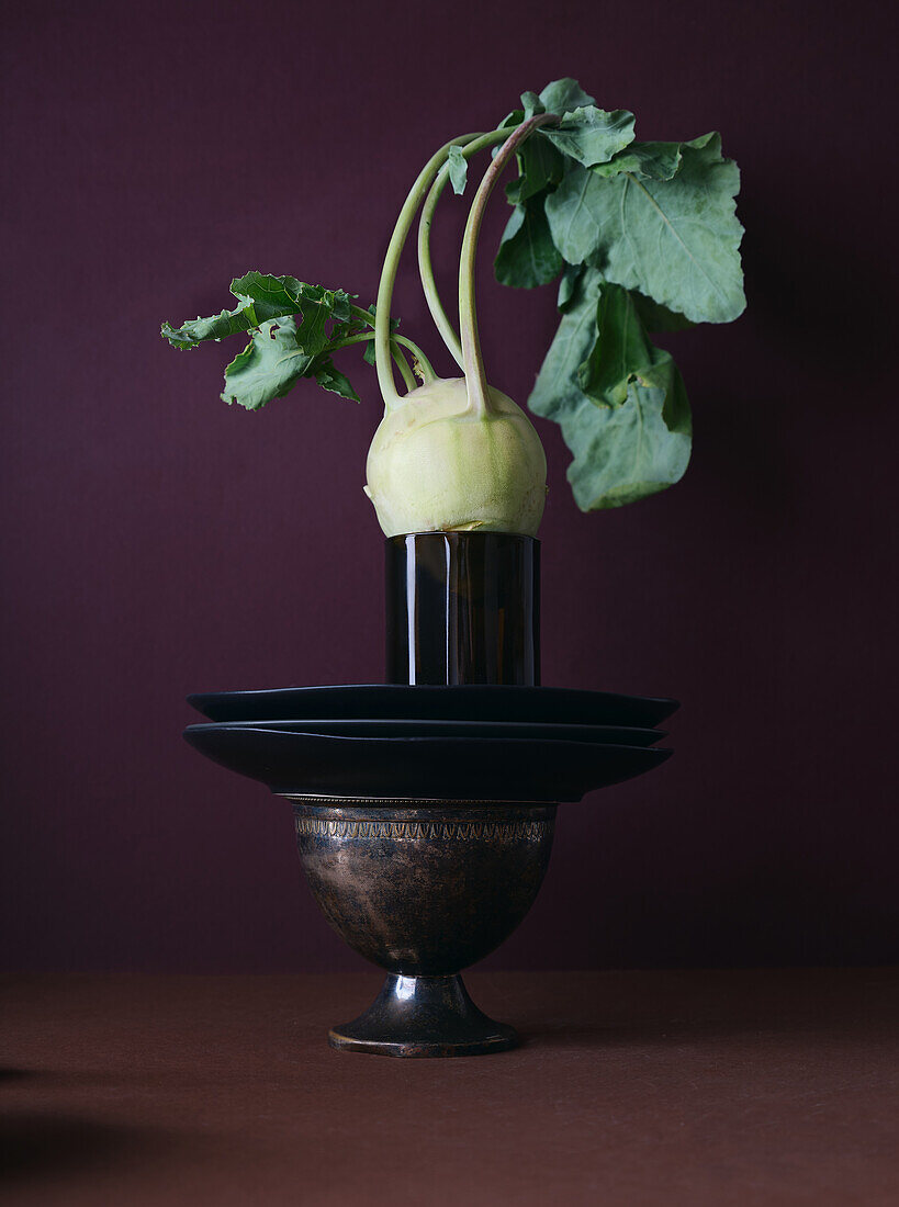 Ein elegant präsentierter Kohlrabi mit frischen Blättern thront auf einem verschnörkelten Vintage-Metallpokal vor einem kastanienbraunen Hintergrund, der eine Mischung aus natürlichen und antiken Elementen darstellt