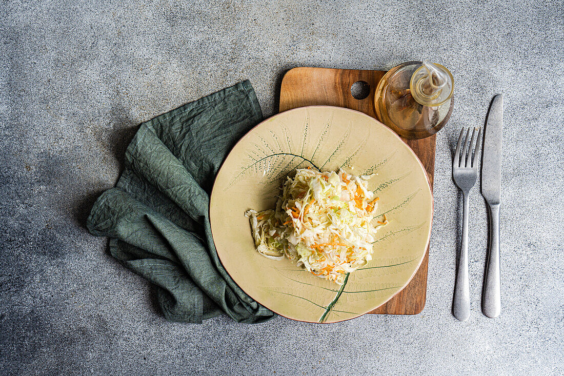 Top view of an organic cabbage and carrot salad drizzled with olive oil, neatly served on a ceramic plate accompanied by a wooden board and silver cutlery, placed on a textured gray surface.