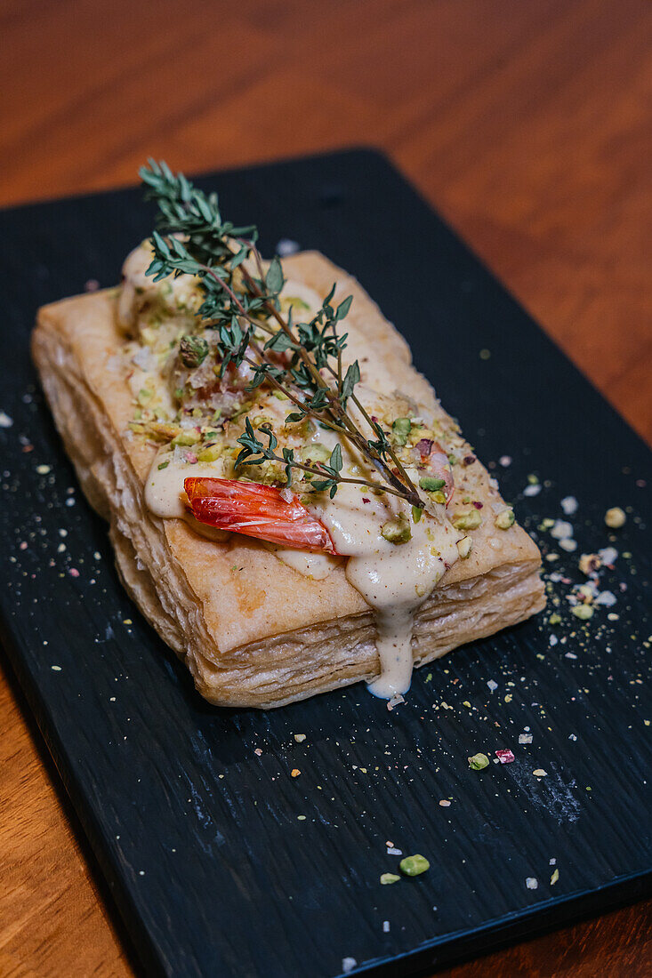 A sumptuous puff pastry dish, garnished with herbs, a succulent shrimp, and drizzled with a creamy sauce, presented on a sleek black slate