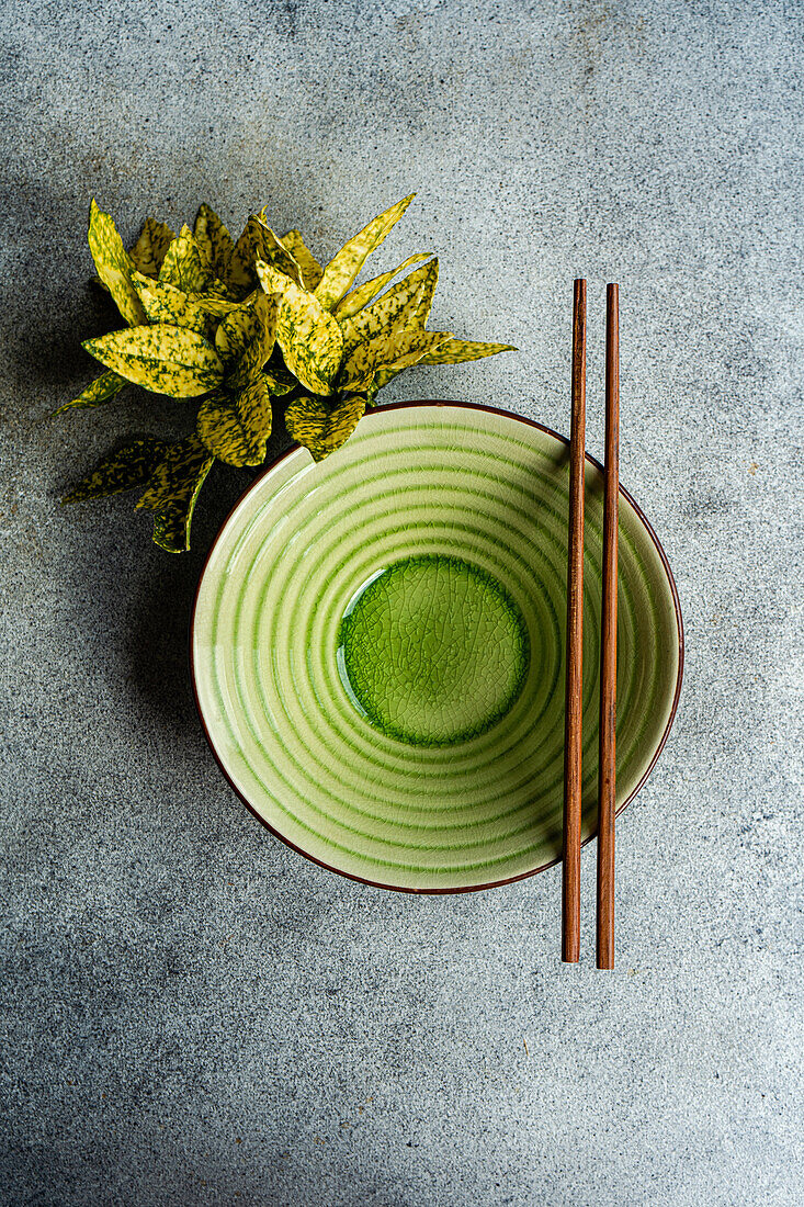 From above view of a neatly set table with a green ceramic bowl and brown chopsticks, accompanied by a potted plant.