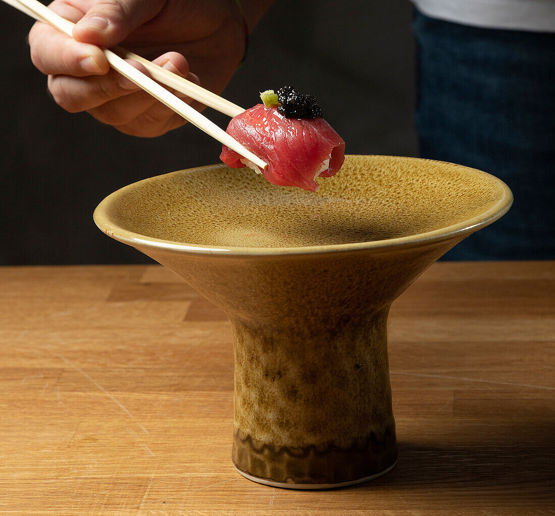 Ein Koch greift mit Stäbchen ein Stück Thunfisch-Nigiri mit Kaviar, das über einer strukturierten Keramikschale schwebt