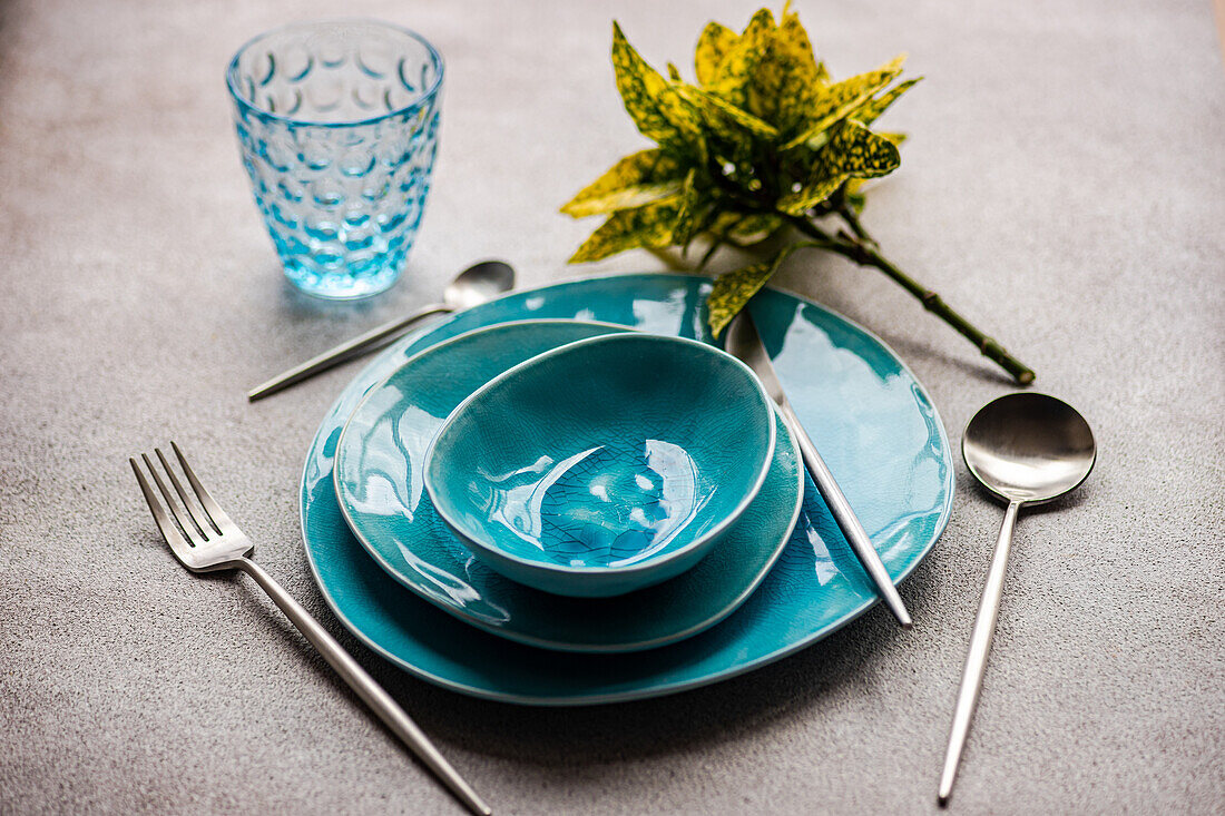 Vibrant teal dinnerware set with matching glass and cutlery on a textured grey surface.