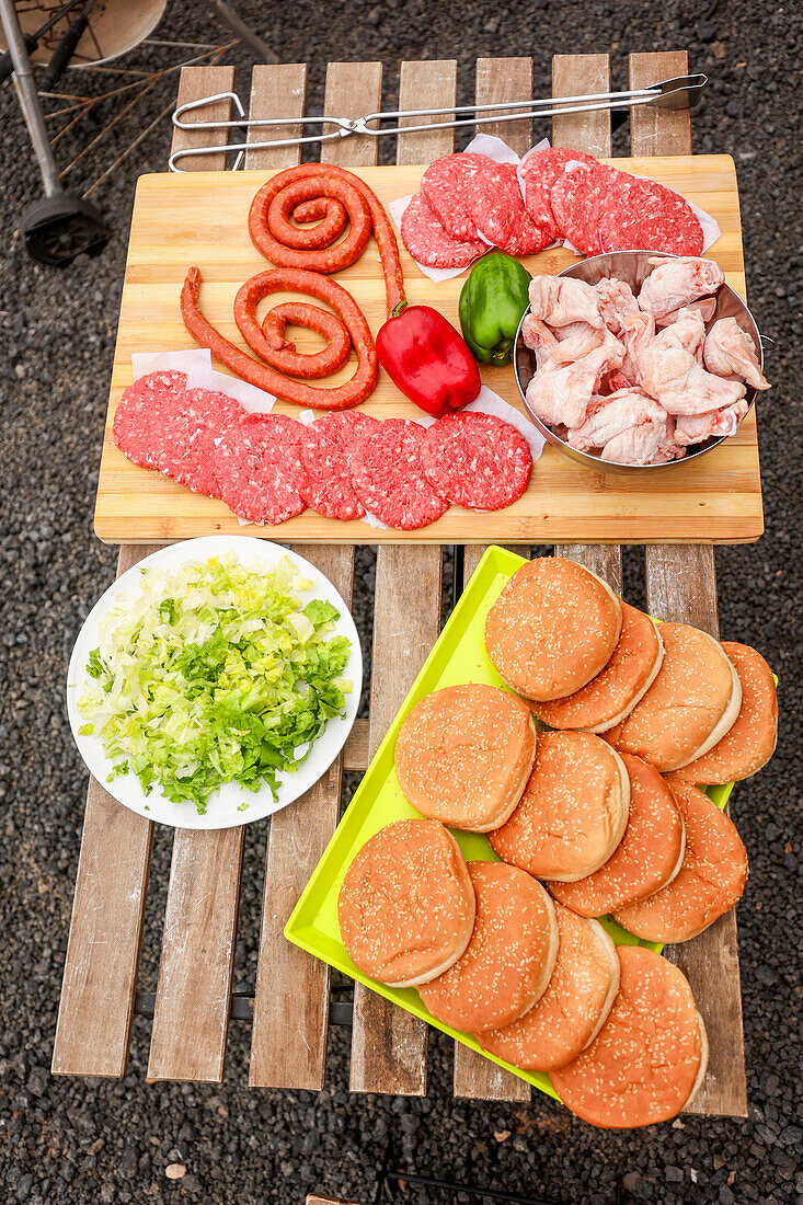 From above top view of an array of barbecue ingredients ready for grilling, including raw sausages, burger patties, chicken pieces, chopped lettuce, and buns displayed on wooden and colorful kitchenware.