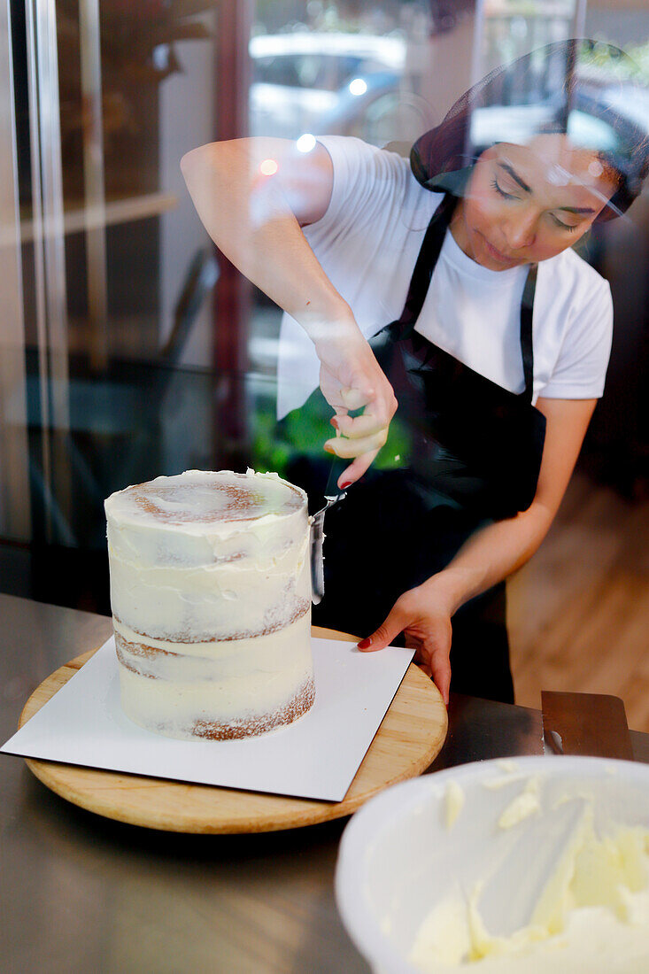 A focused pastry chef is meticulously applying frosting to a multi-layered cake inside a bakery, as seen through a glass window