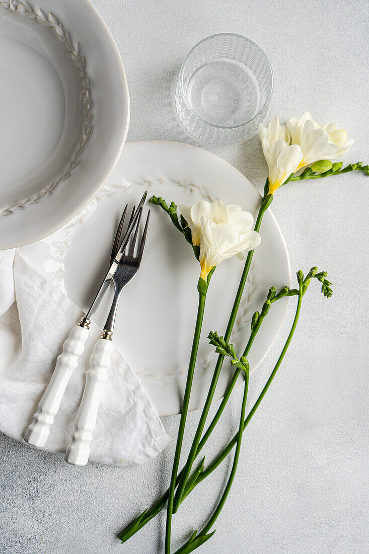 An exquisite table setting featuring delicate white freesia blossoms alongside pristine white dinnerware, fine silverware, and a clear glass The set up is completed on a textured grey backdrop, perfect for a sophisticated dining experience