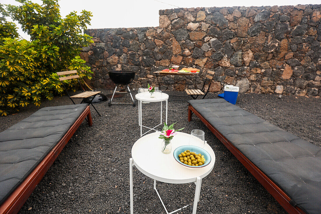 A well prepared outdoor barbecue space featuring a charcoal grill, a table laid with fresh snacks, and a comfortable seating arrangement against a rustic stone wall, all set for a gathering.