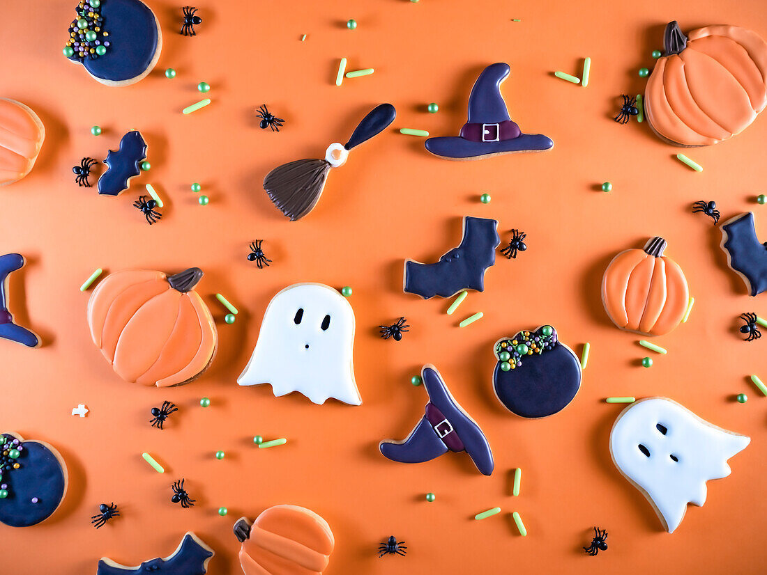 Eine bunte Sammlung selbstgebackener Halloween-Kekse in Form von Geistern, Kürbissen und Hexenhüten, kreativ dekoriert und arrangiert auf einem leuchtend orangefarbenen Hintergrund