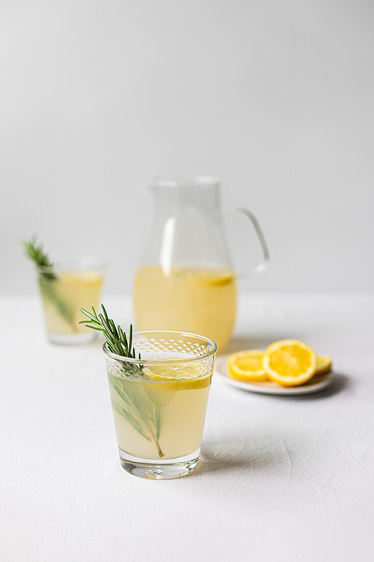 Frische hausgemachte Limonade mit Rosmarin, serviert in einem Glas mit einem Krug im Hintergrund auf einer weißen Fläche